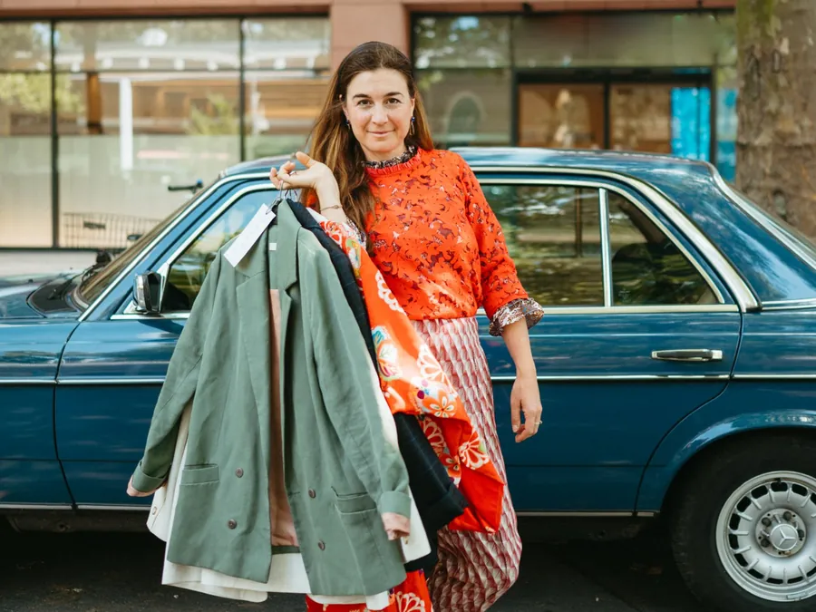 Stylistin Janine Dudenhöffer mit einer Auswahl an Jacken auf Bügeln vor einem blauen Oldtimer